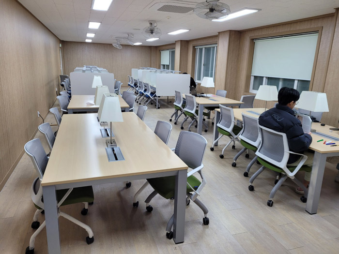 6인용 책상을 가운데 두고 의자가 서로 마주보는 형태를 갖춘 모습의 책상과 의자가 4세트 놓여있다.